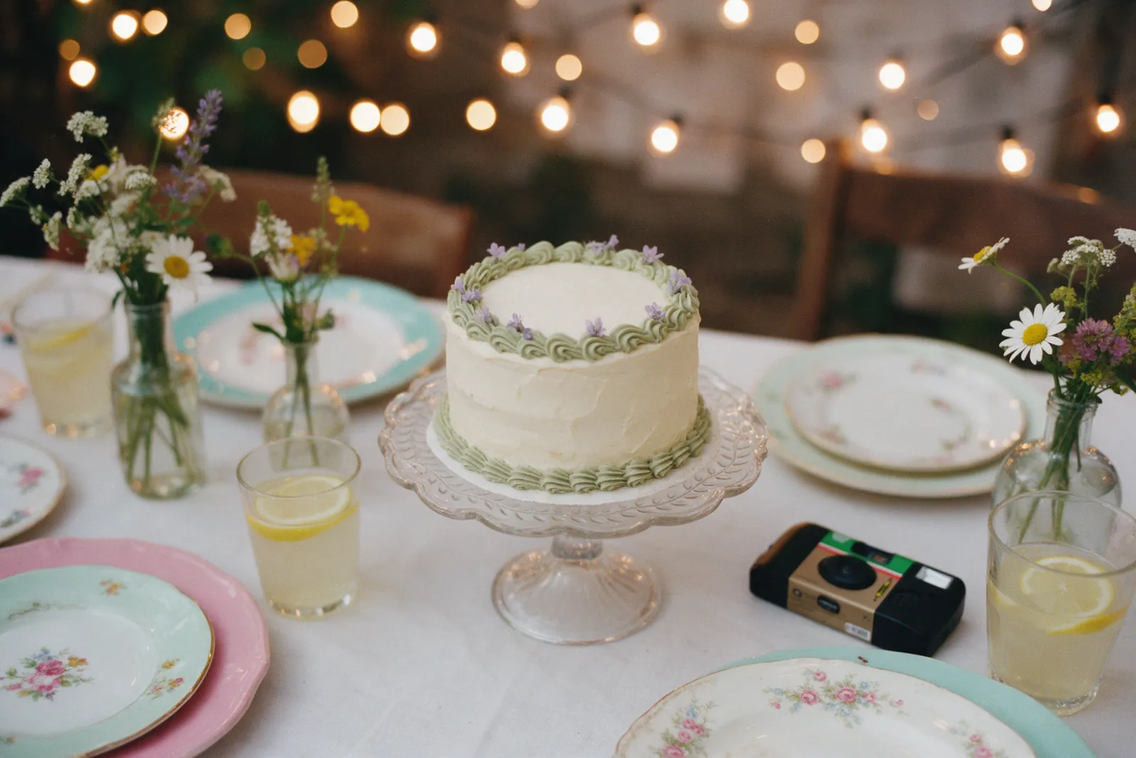 A small cream cake with sage green piping on a glass stand, surrounded by lemonade and wildflowers.