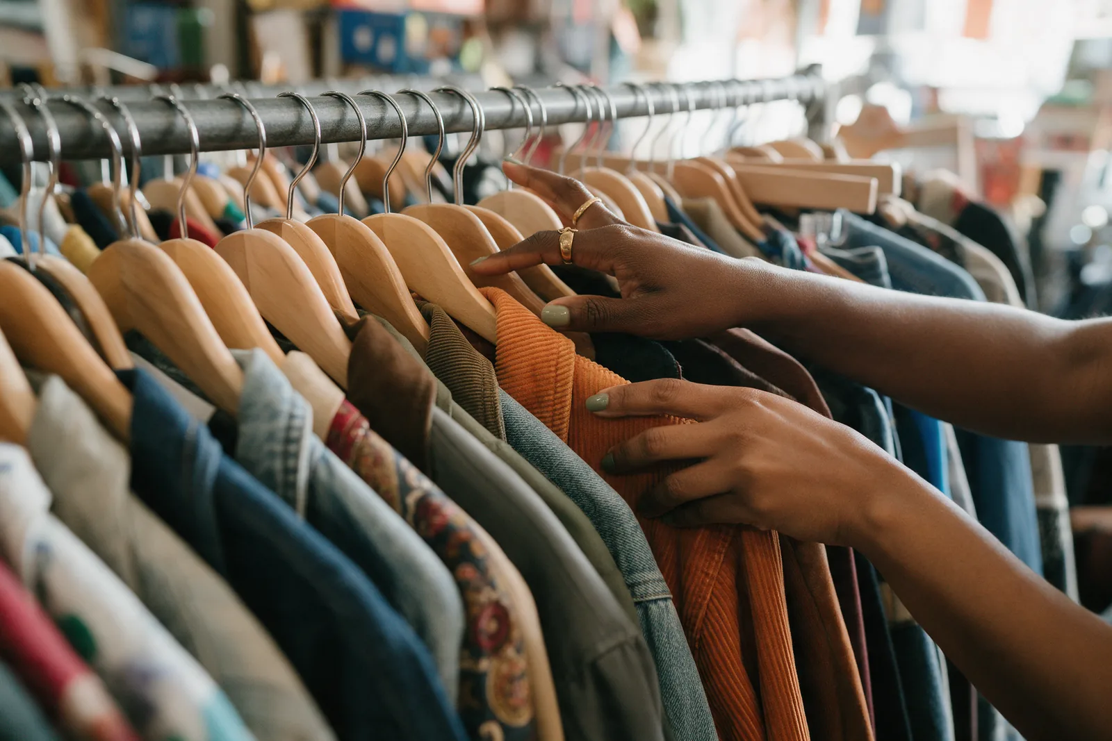 Hands sliding hangers along a crowded thrift rail, pausing on an orange corduroy jacket.